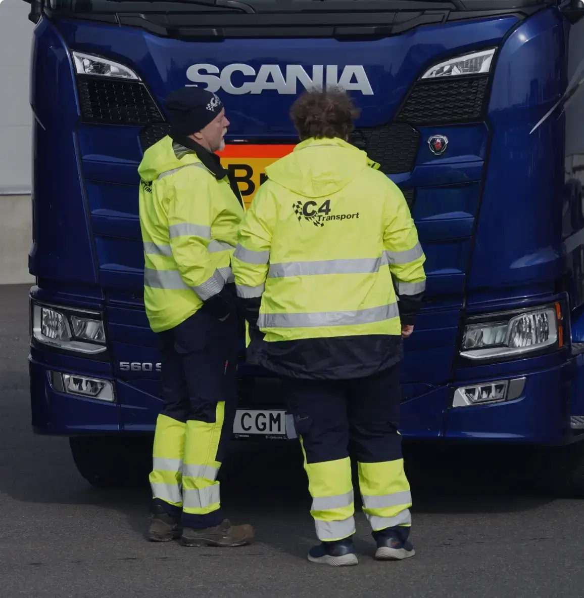Två personer i gula skyddsjackor står framför en blå Scania-lastbil.