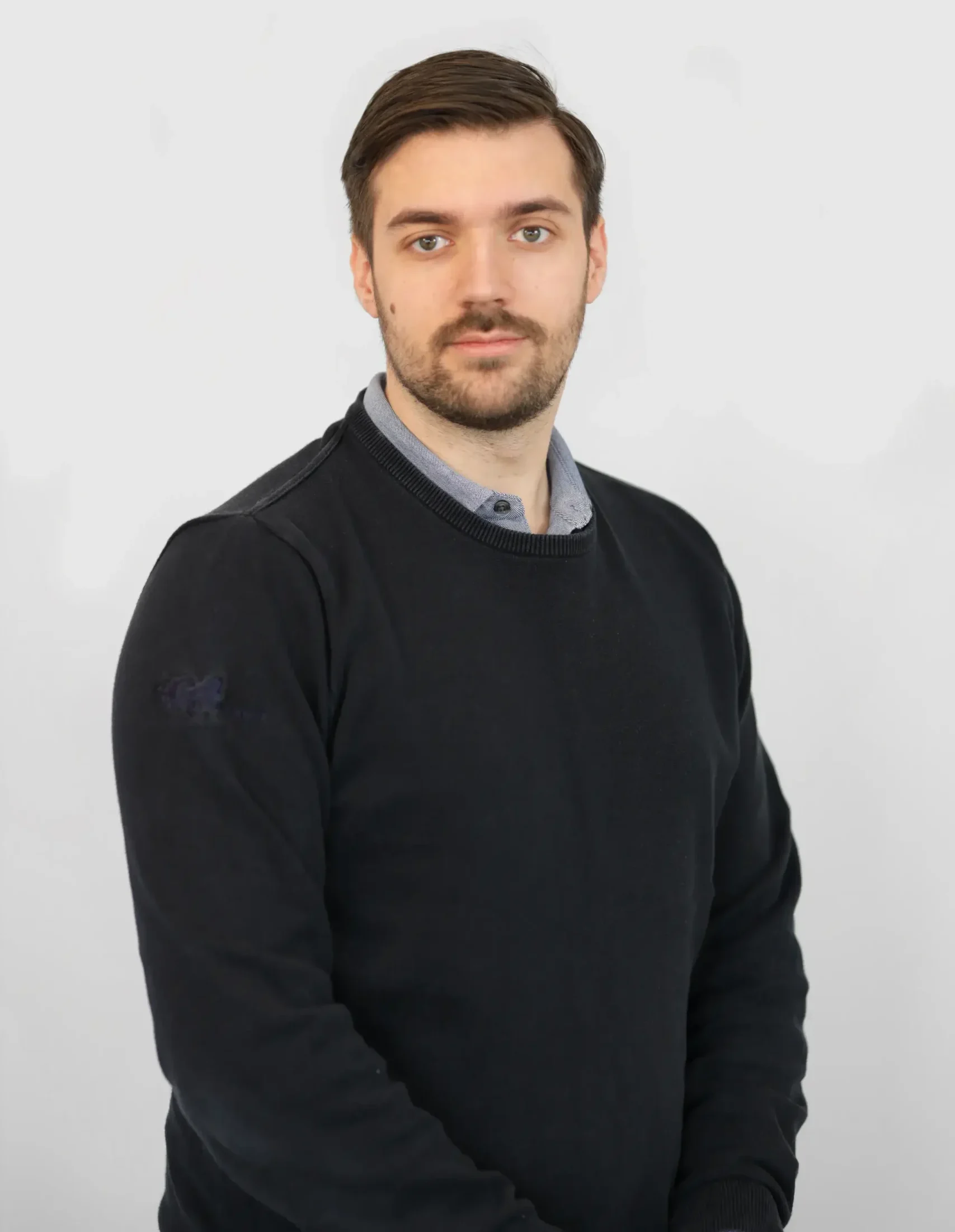 En man med brunt hår och skägg iklädd en svart tröja poserar mot en ljus bakgrund.