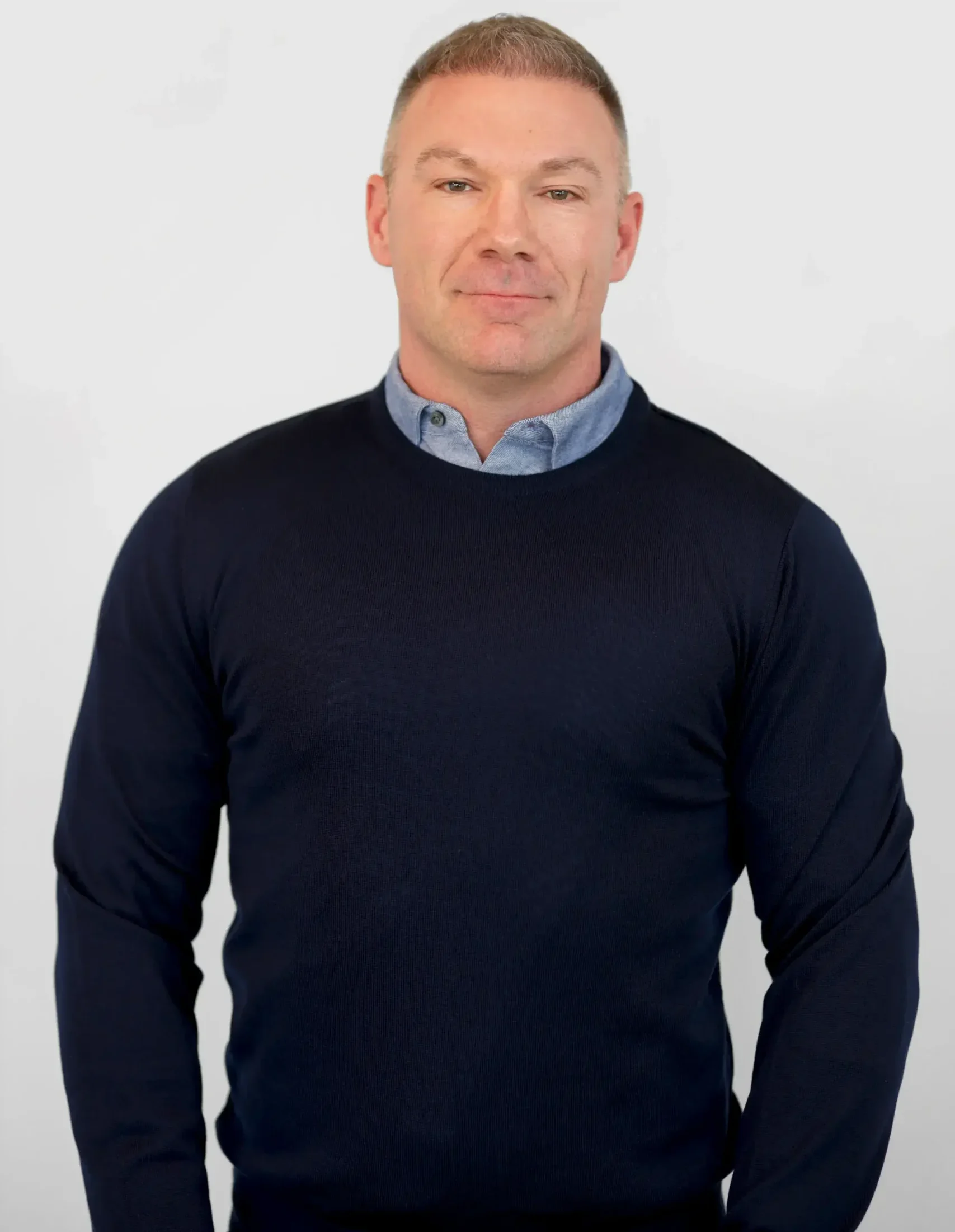 Man i en marin tröja och skjorta med blå krage stående mot en vanlig ljus bakgrund.