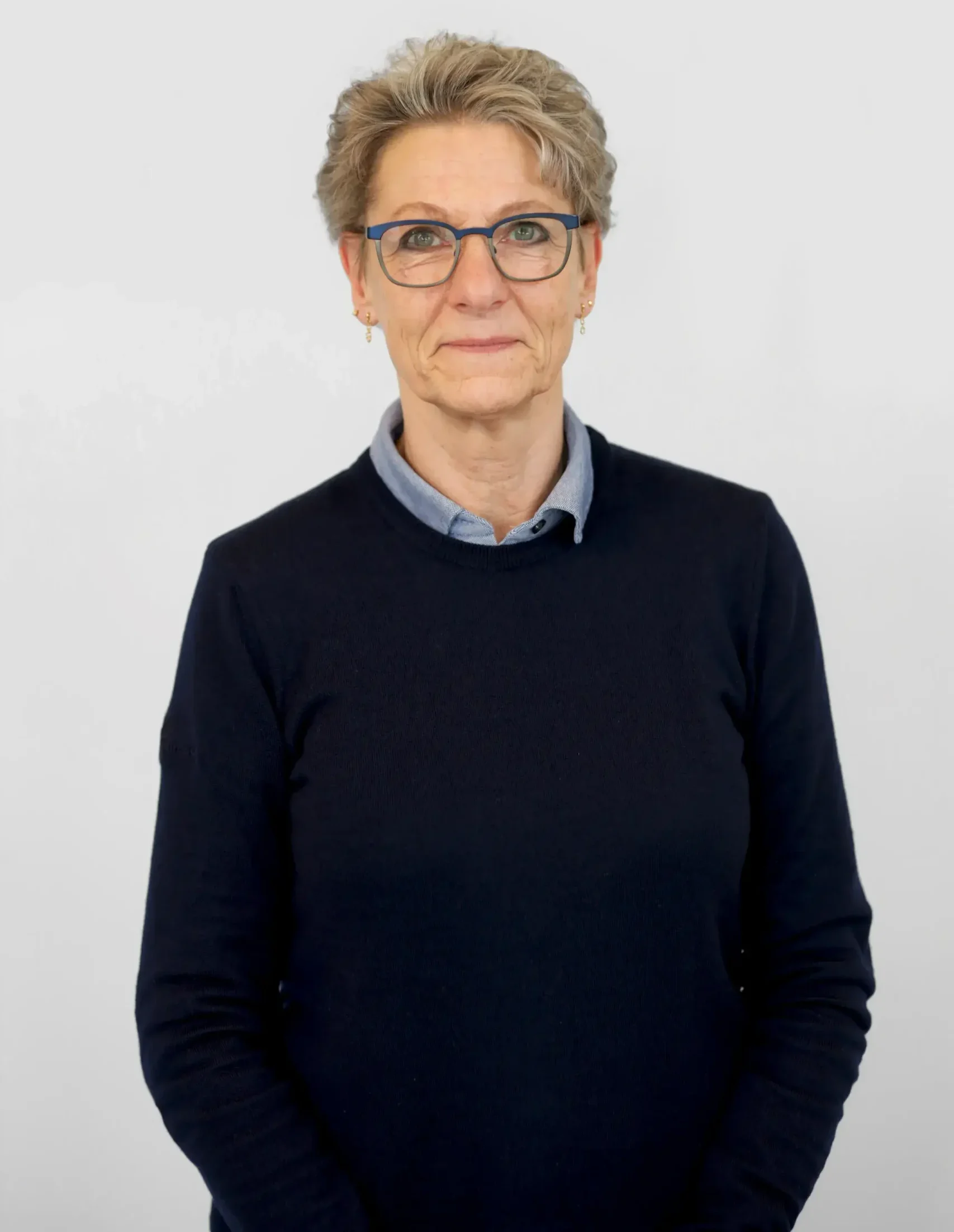 Äldre person med kort hår, glasögon och mörk tröja som står mot en vanlig bakgrund.