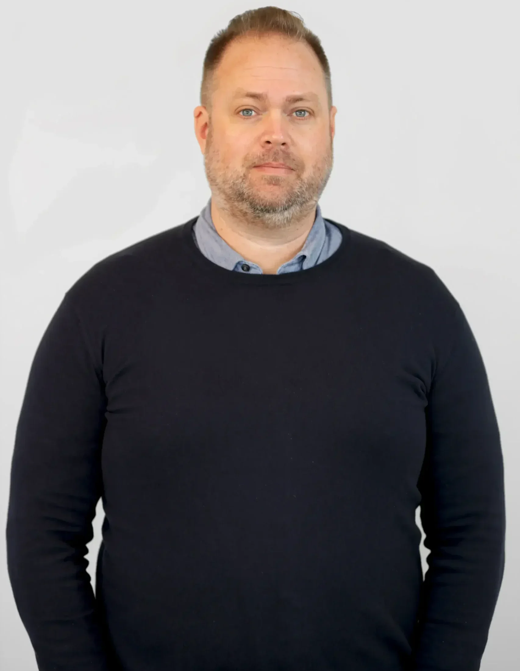 Man med kort hår och skägg klädd i en mörk tröja, stående mot en vanlig bakgrund.