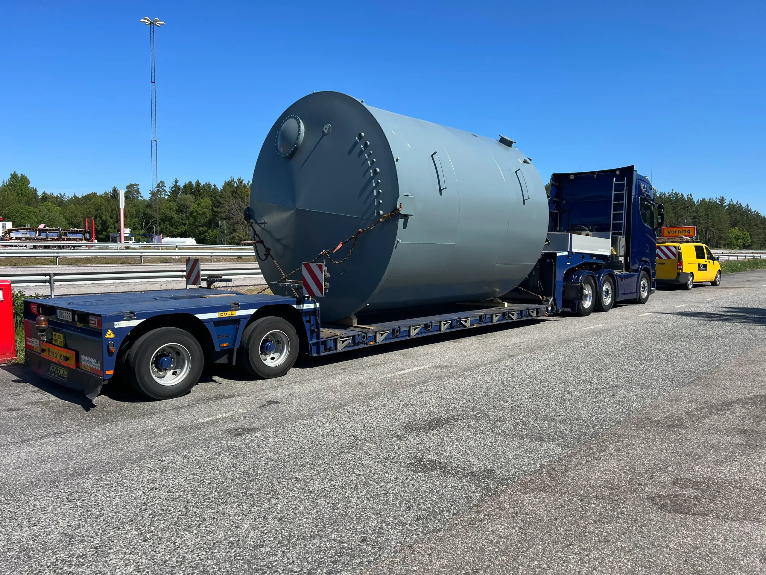 Stor cylindrisk tank lastad på en flakvagn, parkerad bredvid en väg en solig dag.
