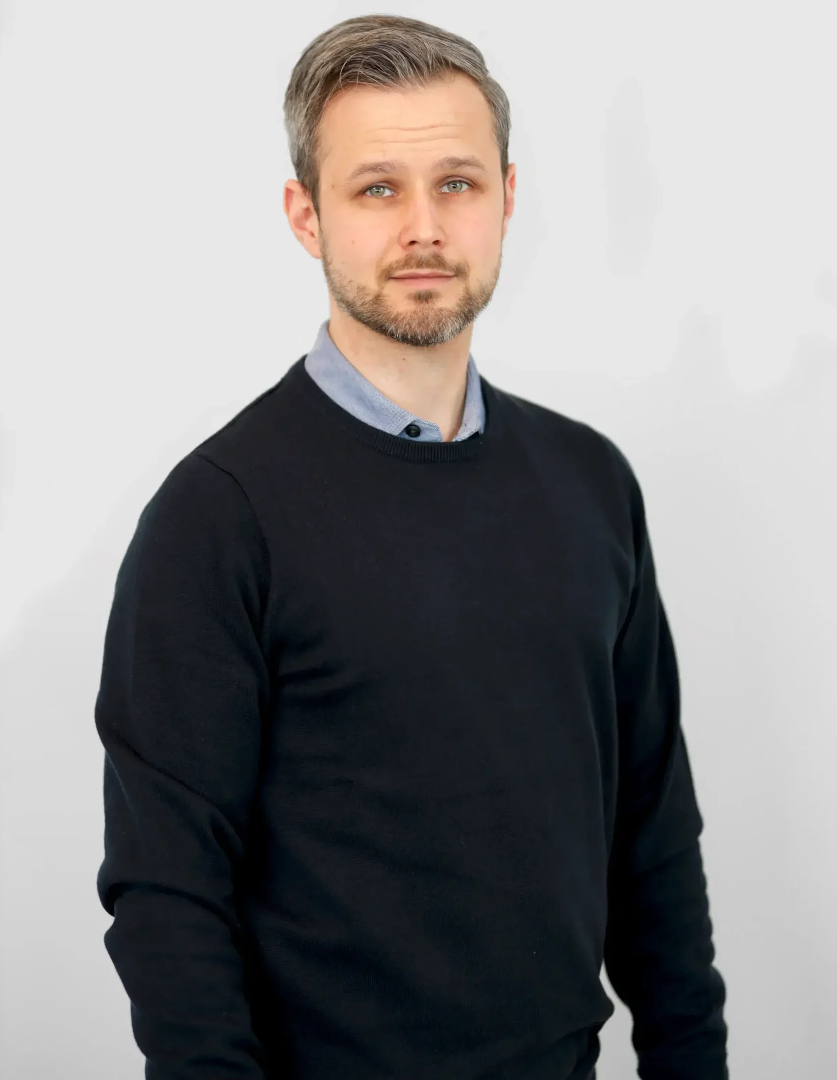 Man med kort hår och skägg bär en svart tröja över en krage skjorta, stående.