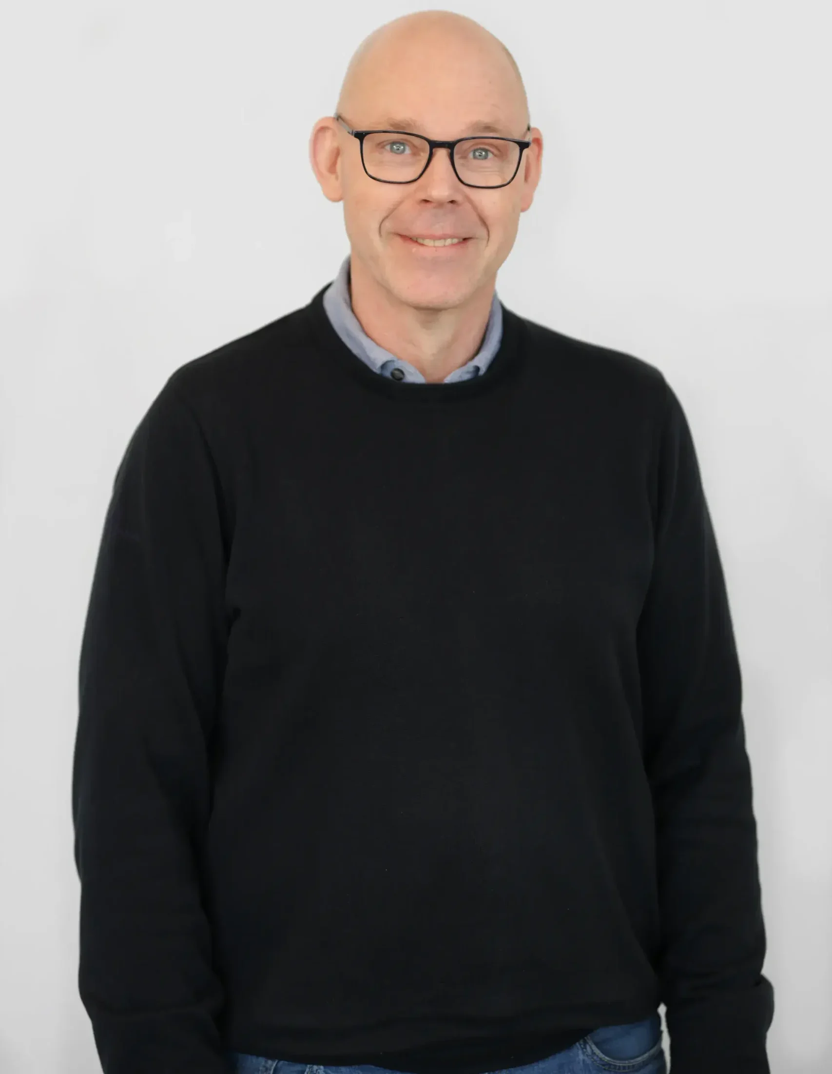 Skallig man med glasögon i en svart tröja ler mot kameran mot en vanlig bakgrund.