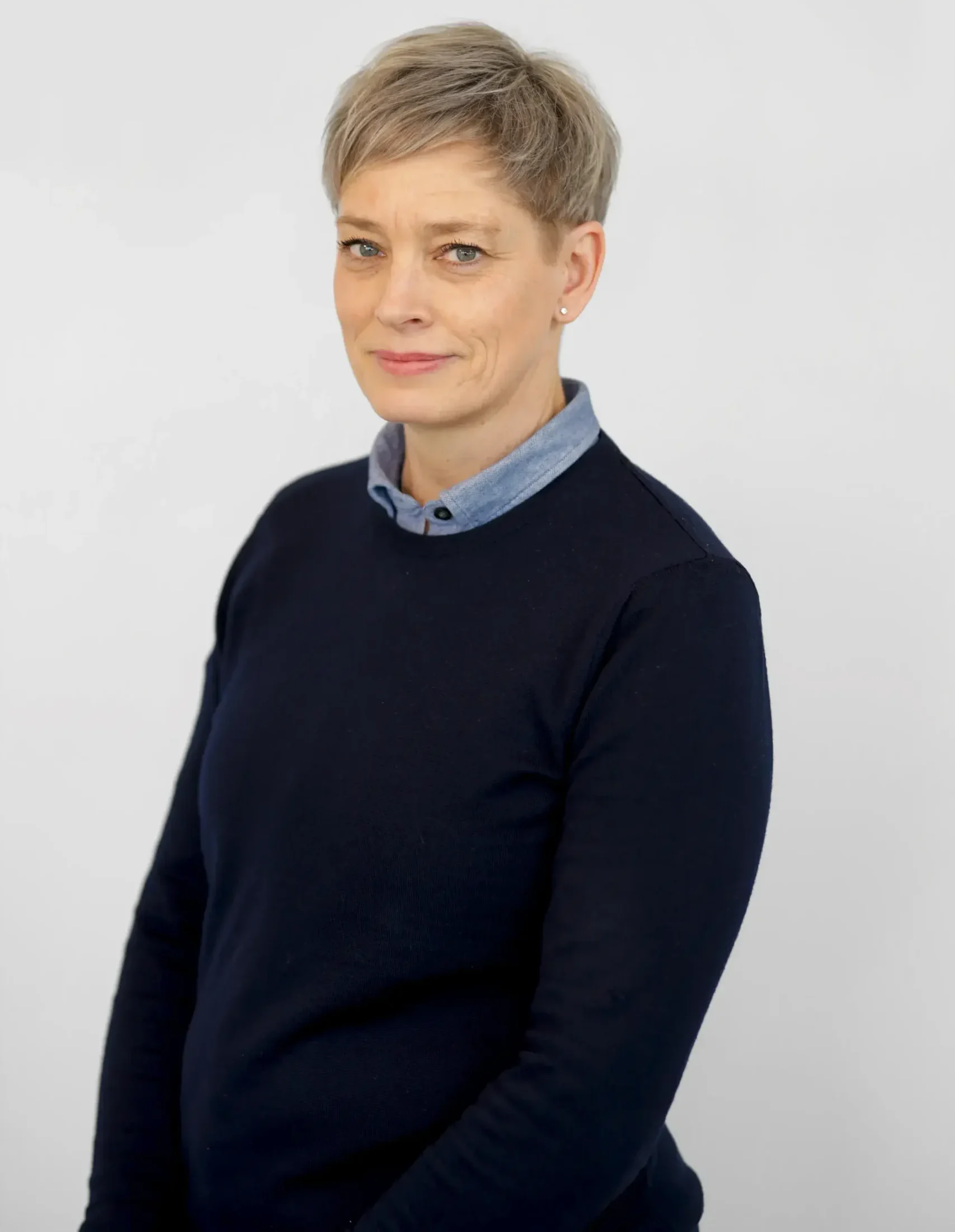 Person med kort blont hår i en marin tröja och skjorta med krage poserar mot en ljus bakgrund.
