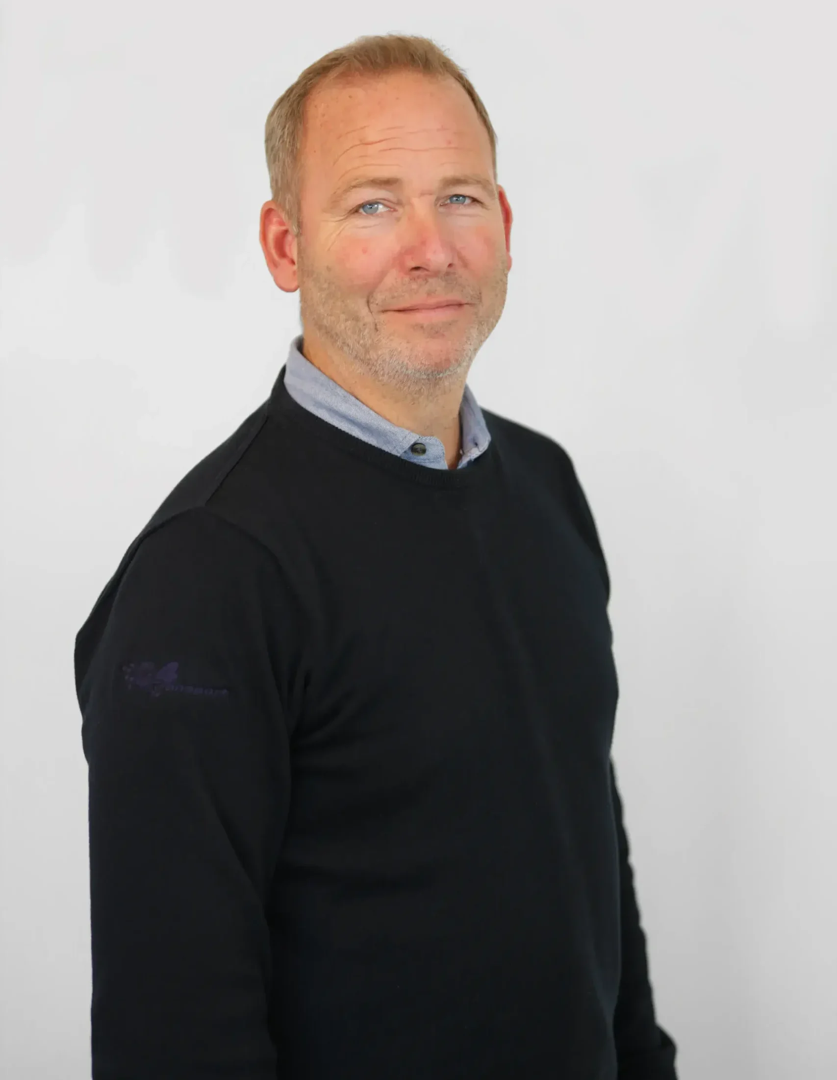 Man i en svart tröja och skjorta med krage stående mot en vanlig ljus bakgrund.